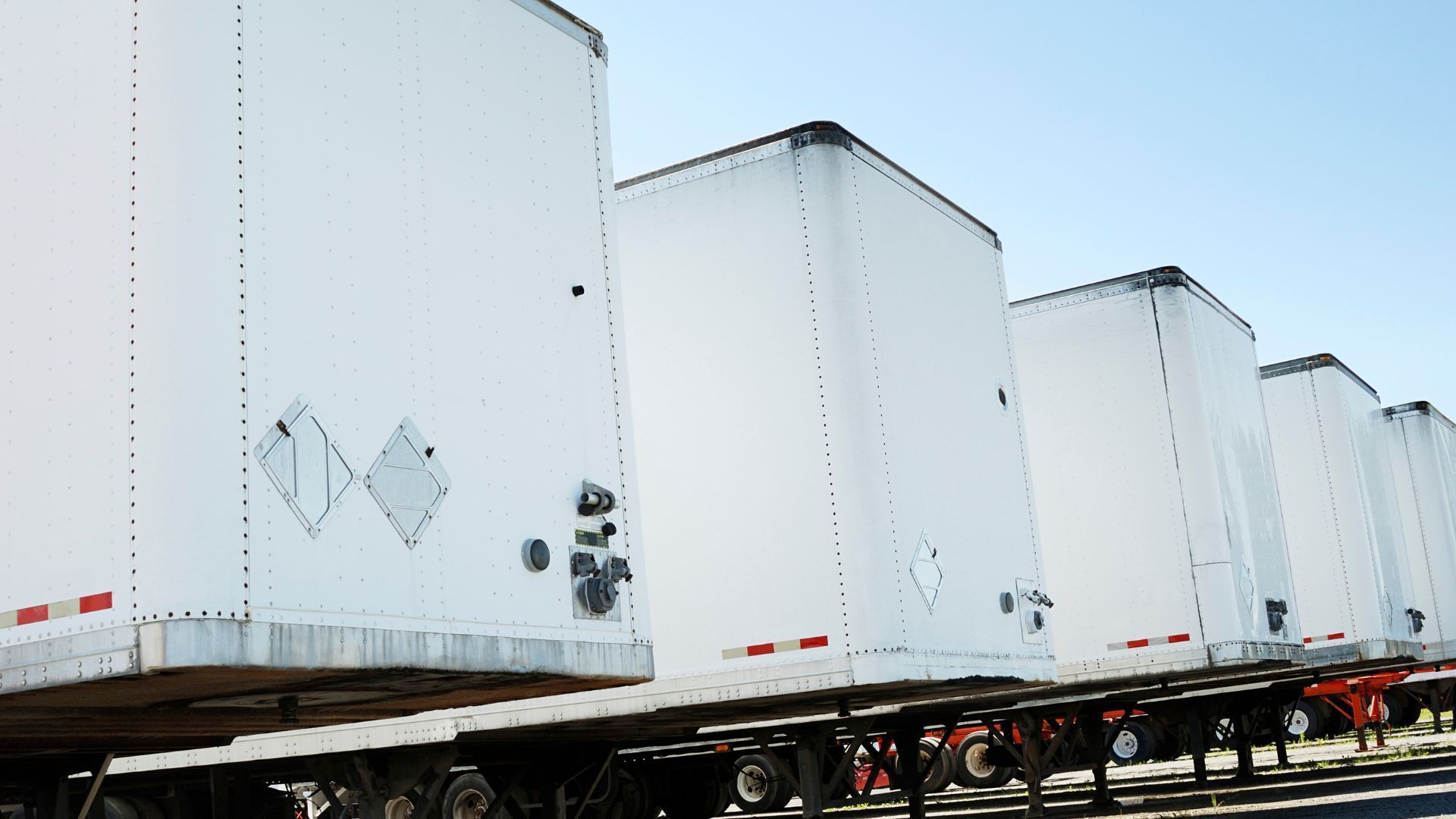 White semi-trailers lined up in a row on a highway or lot against a pale sky.