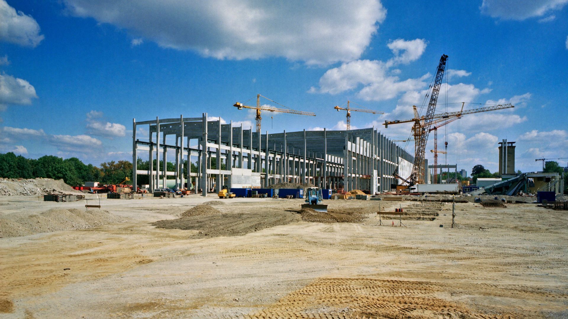 Construction site with steel building frame, cranes, and dirt foreground under a blue sky