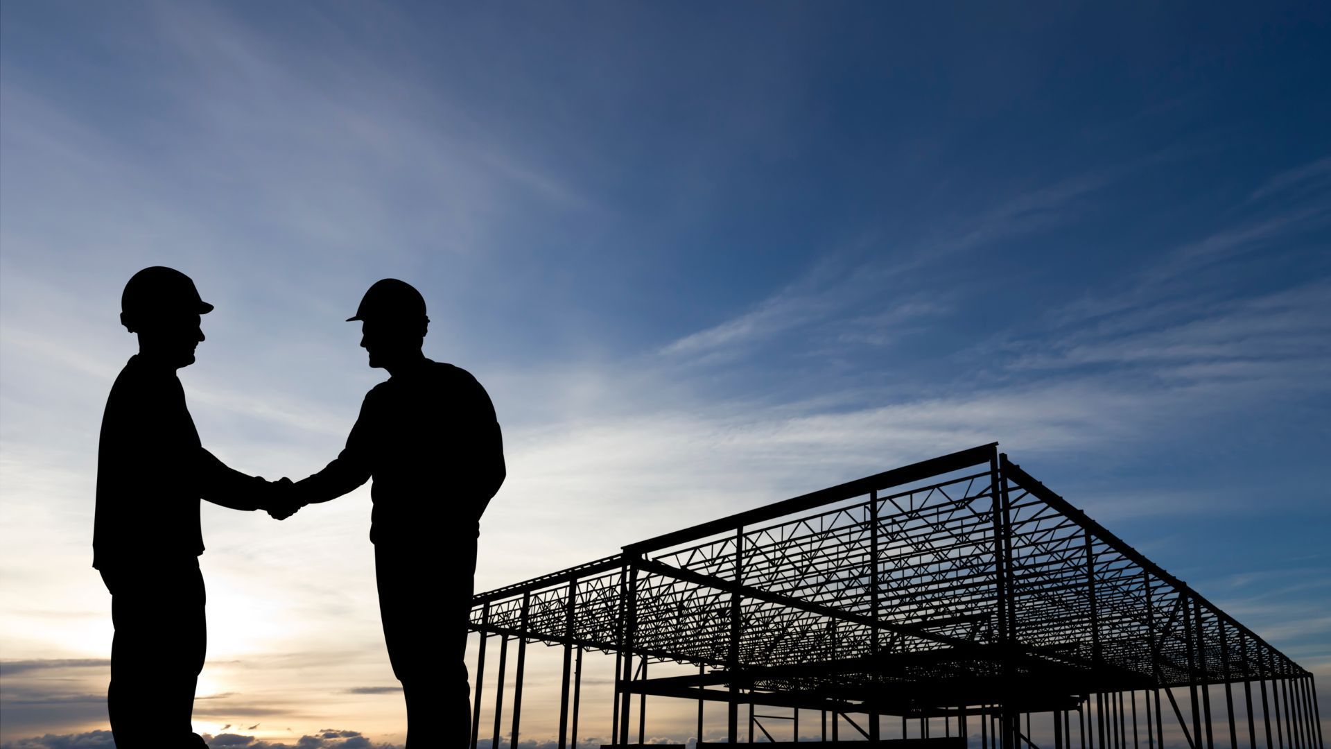 Two workers in hard hats shake hands at a construction site at dusk.