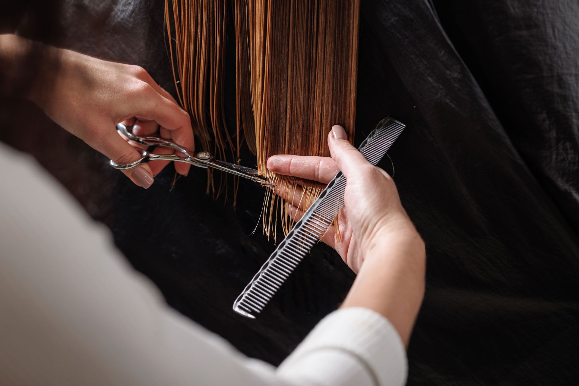 Hands cutting wet, light-brown hair with scissors and a comb against a black backdrop.