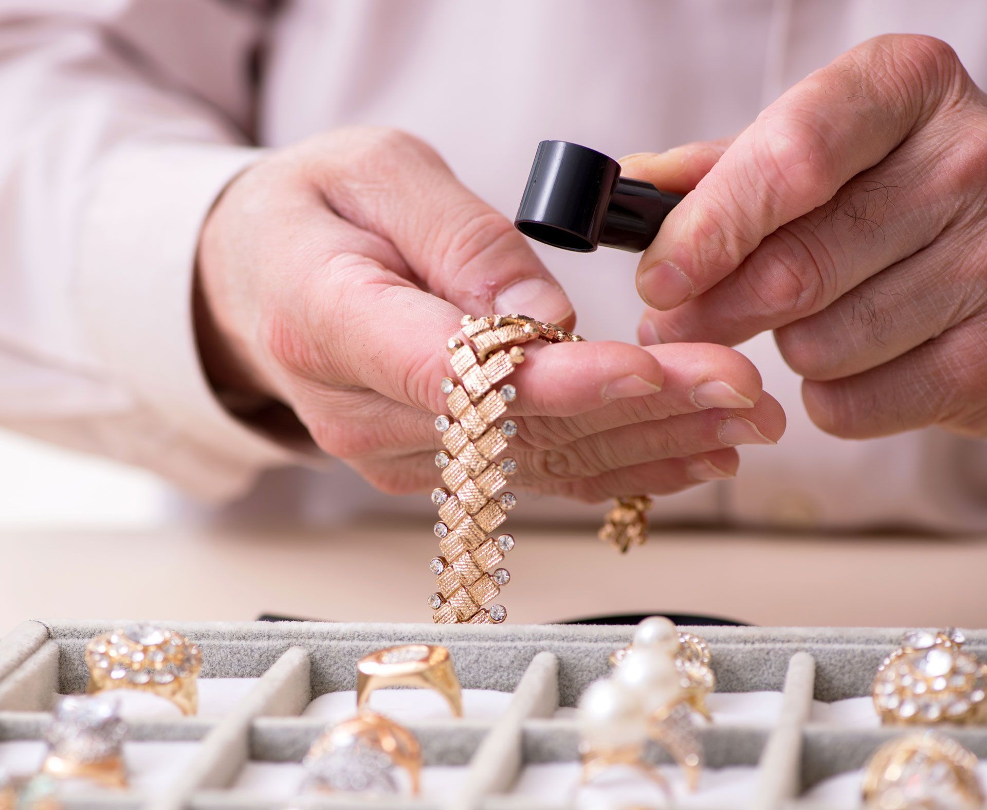 Person examining a gold bracelet with a jeweler's loupe, with jewelry display case in the foreground.