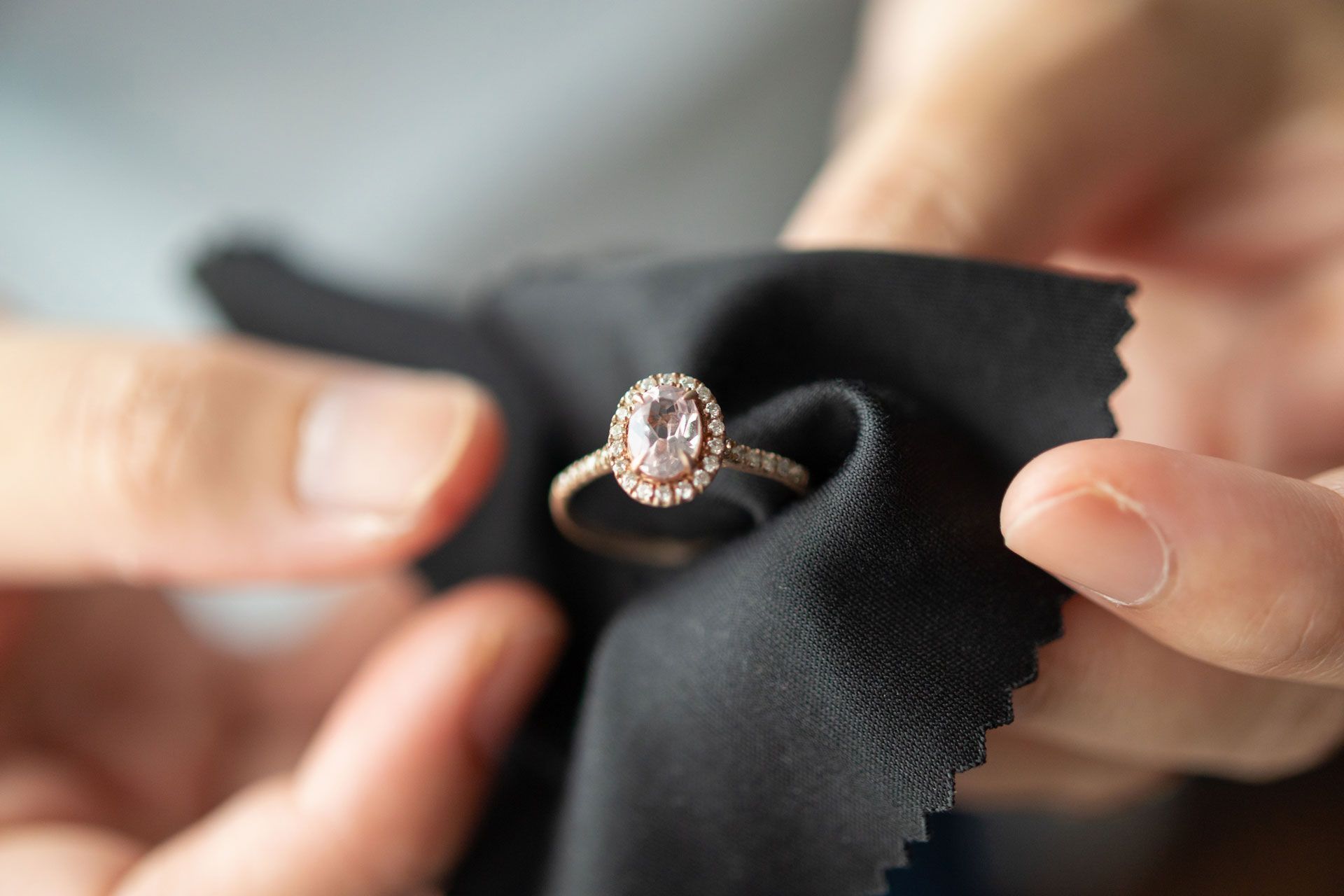 Hands holding a gold ring with a pink stone and diamond halo, wiping it with a black cloth.