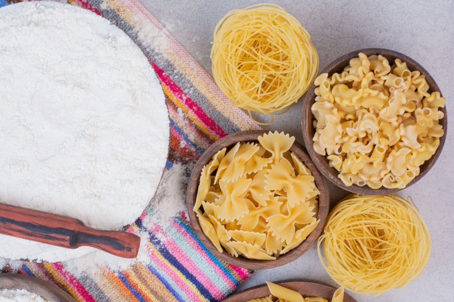Cuencos con pastas de diferentes formas, manojos de capellini y un rodillo sobre un paño a rayas espolvoreado con harina.