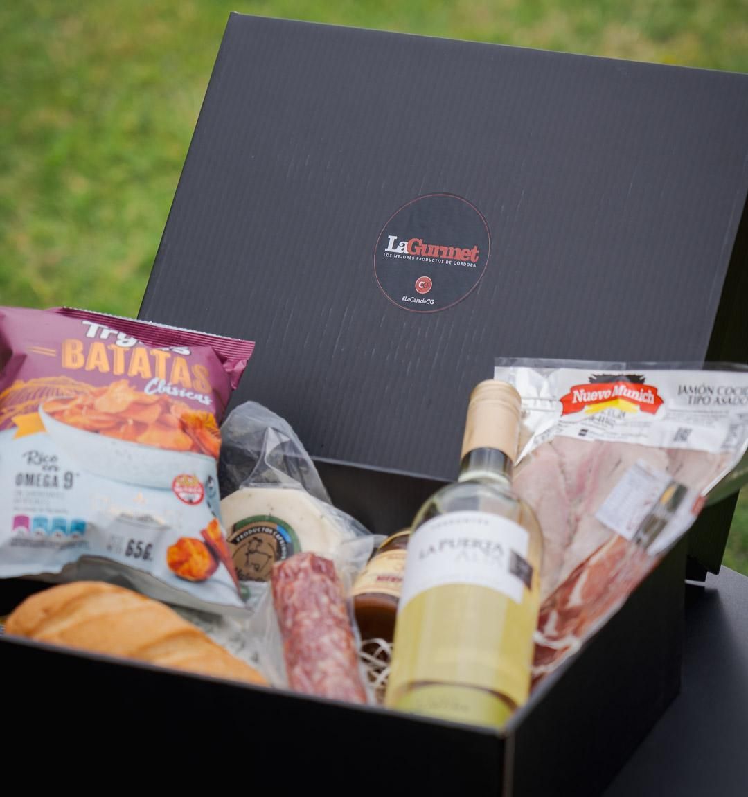 caja de regalo negra que contiene aperitivos, salchicha, vino y un paquete de jamón, colocada al aire libre sobre el césped.