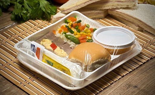 bandeja de comida envuelta en plástico con un panecillo, verduras , taza pequeña y condimentos sobre una esterilla de bambú.
