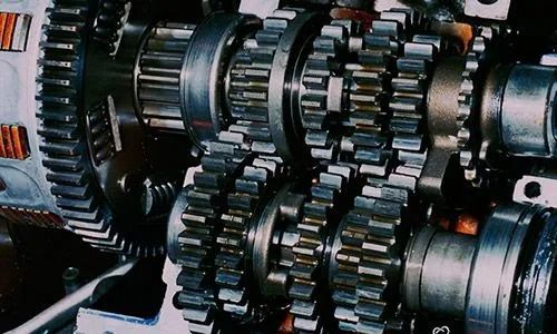 Primer plano de engranajes y ejes metálicos entrelazados dentro de una máquina, con componentes oscuros y plateados.