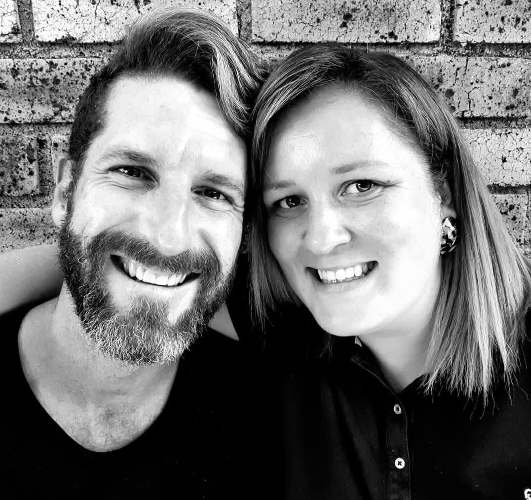 Smiling couple posing close together in front of a brick wall, in black and white.