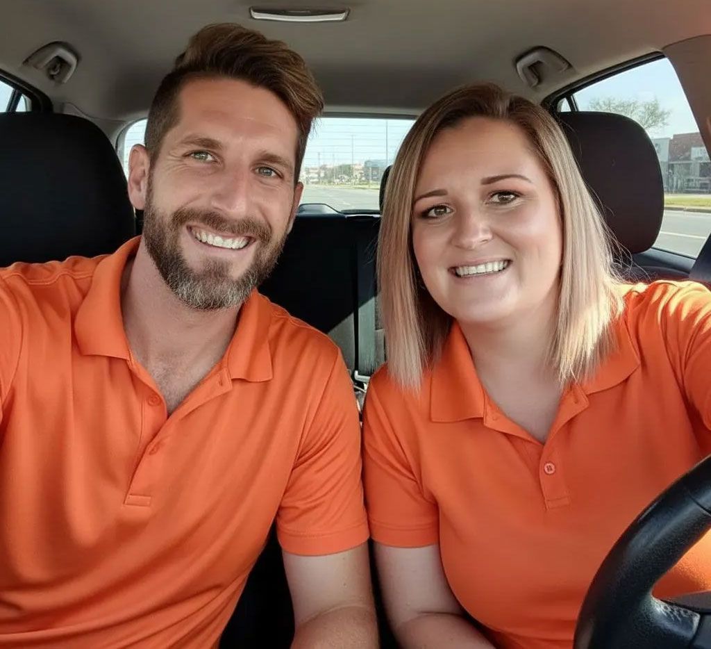 Two smiling people in orange shirts sit inside a car, facing the camera.