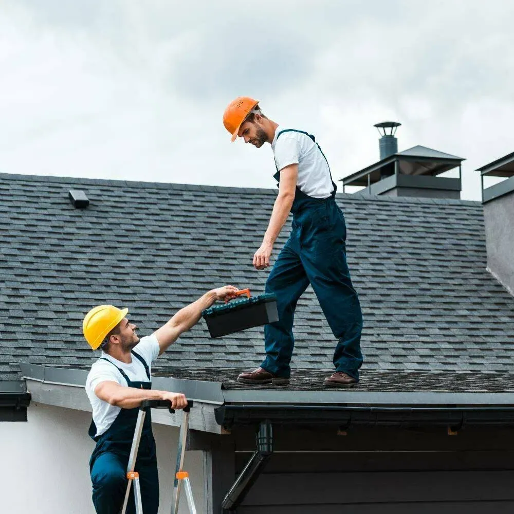 Two roof workers in hard hats passing shingles on a gray shingled rooftop