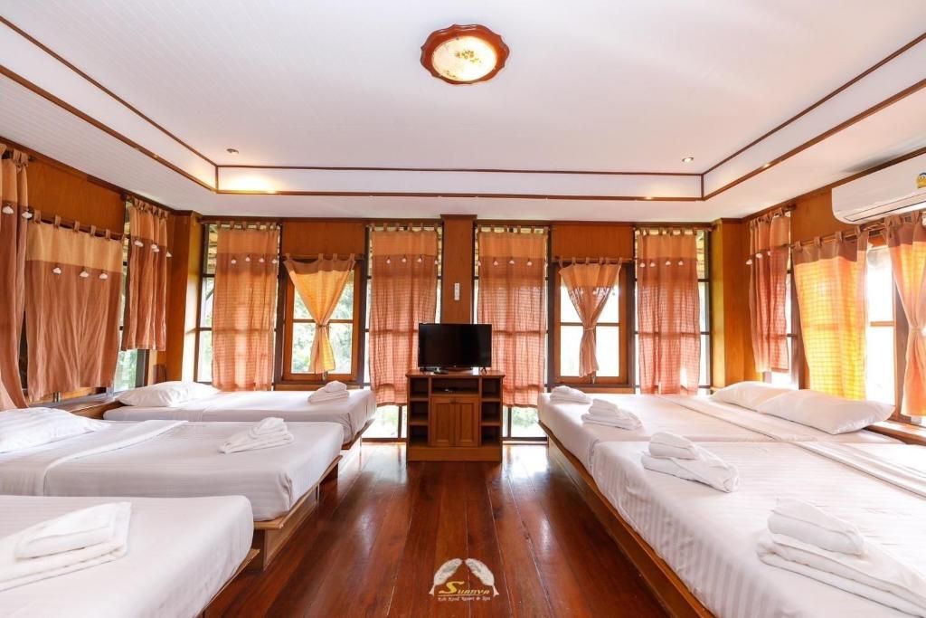 A room with several beds, windows with curtains, and a TV. Wooden floors and walls are visible.