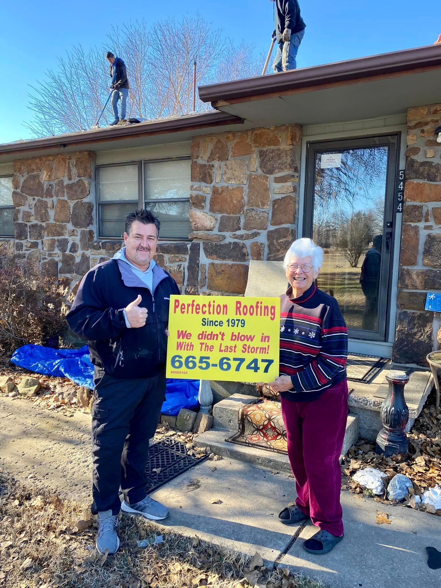 A man and a woman are standing in front of a stone house holding a sign that says perfection roofing