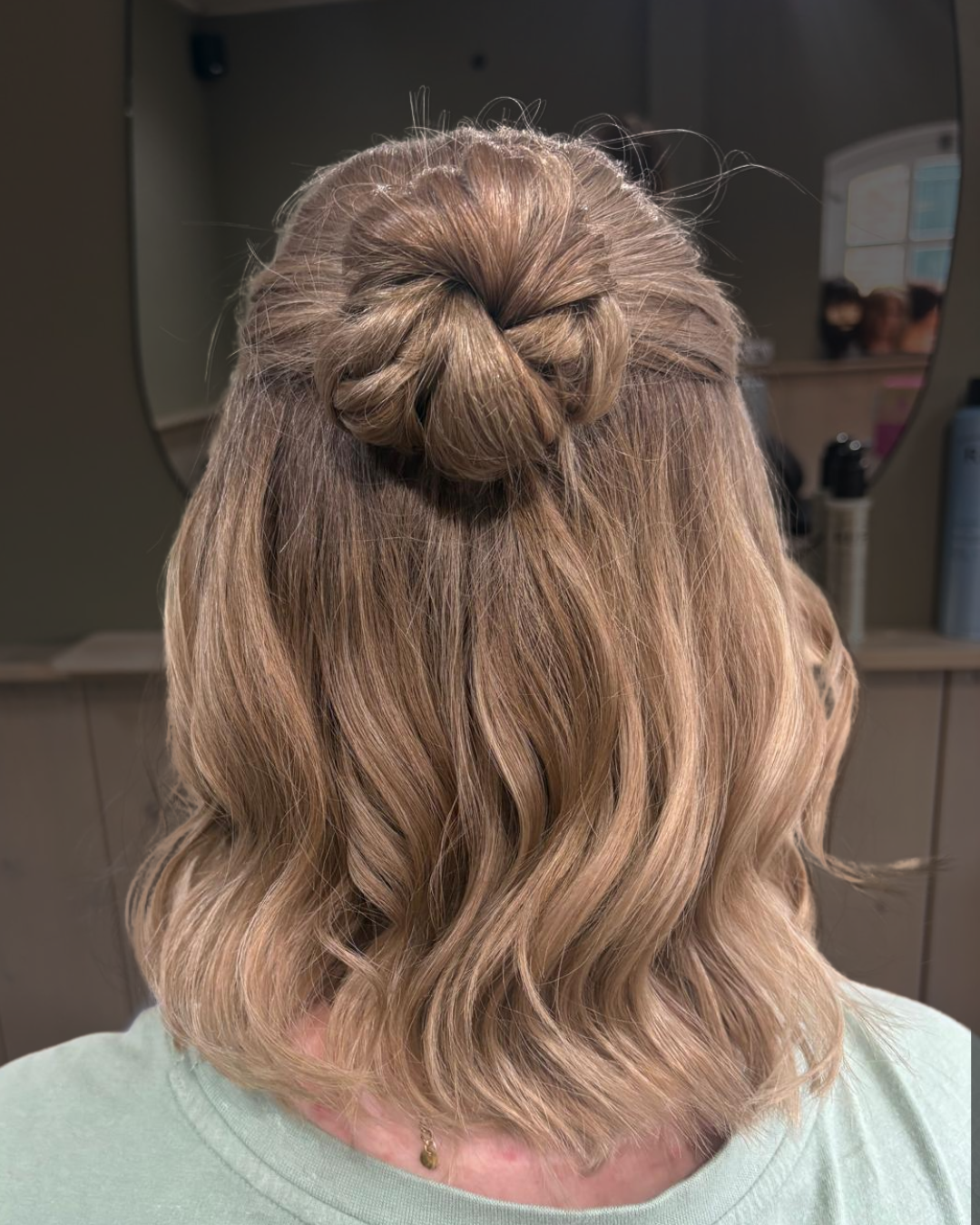 Achteraanzicht van golvend blond haar dat half opgestoken is in een gevlochten knot, met een lichtgroene top aan, binnen.