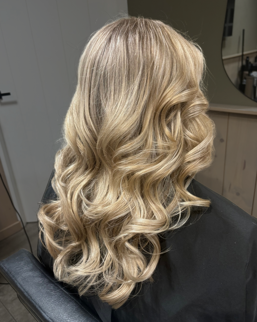 Blond, golvend schouderlang kapsel, van achteren gezien terwijl de vrouw in een kappersstoel zit.