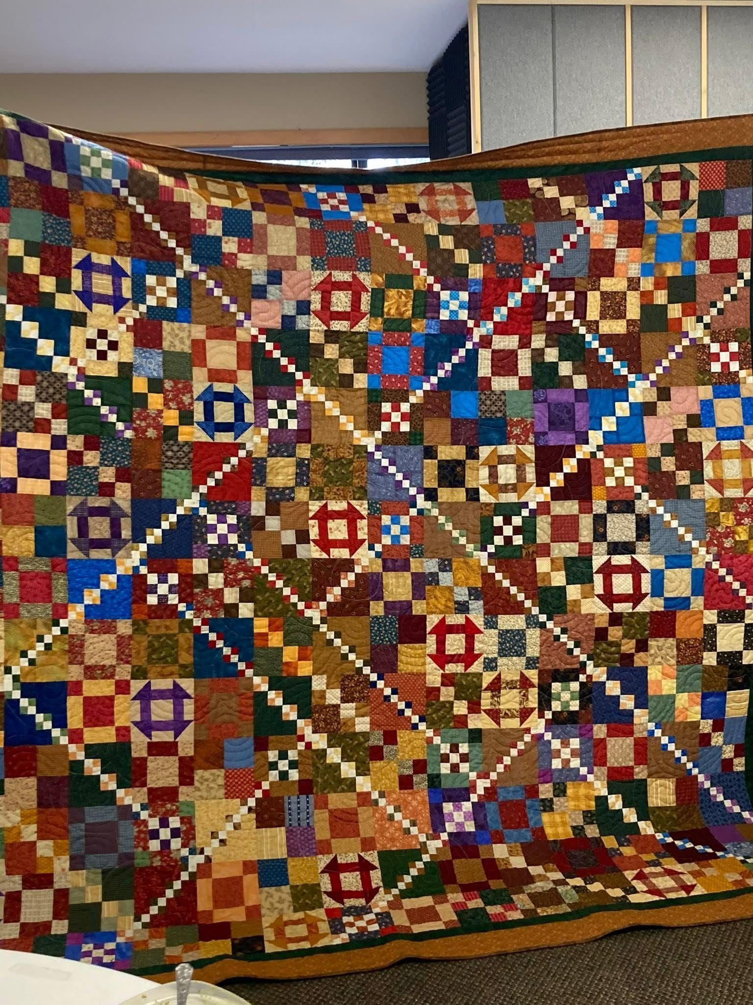 A colorful quilt featuring a patchwork grid pattern intersected by diagonal white and colored lattice chains.