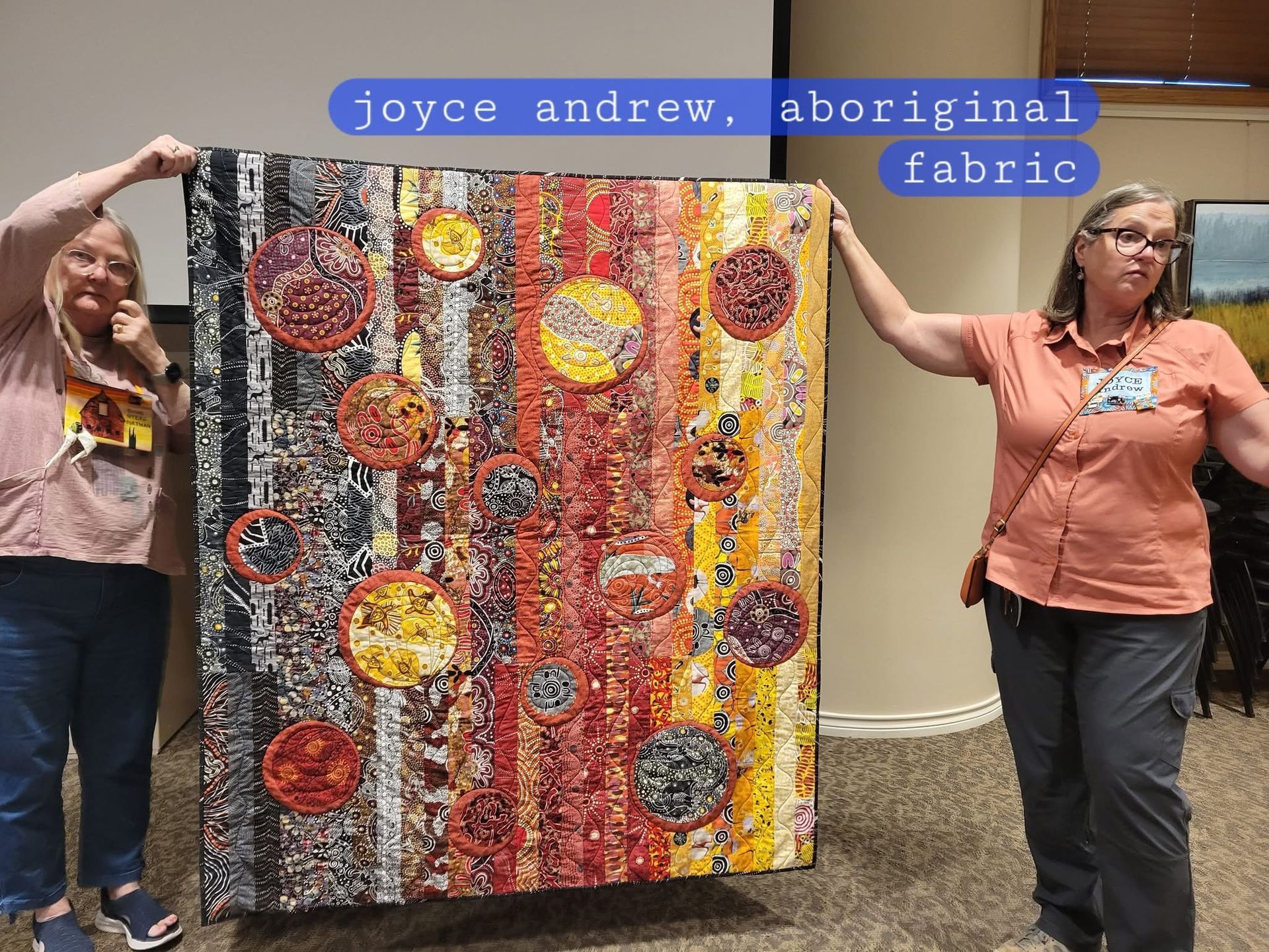Two people hold up a large, colorful quilt featuring vertical fabric strips and circular Aboriginal art patterns.