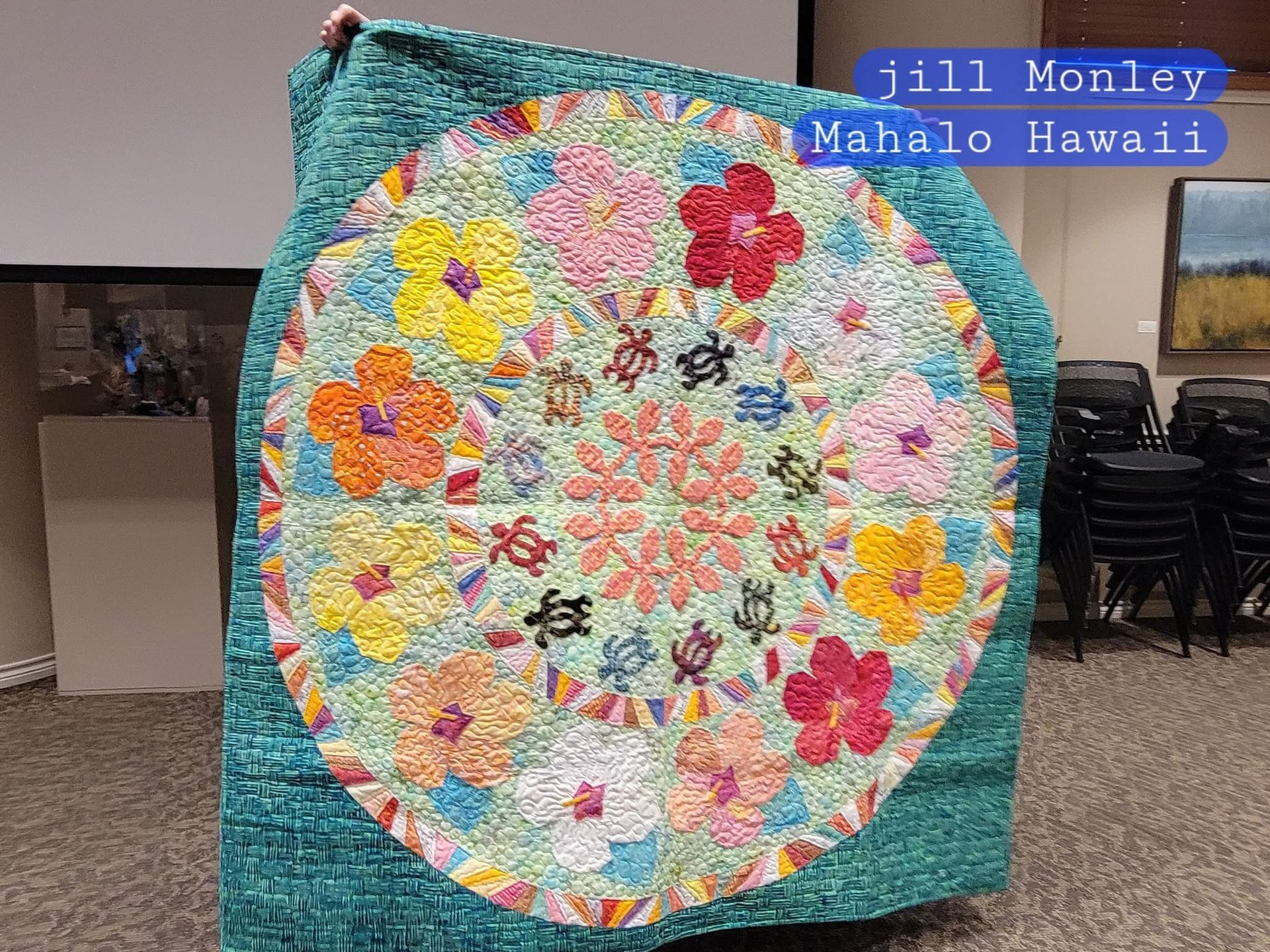 A person holds up a circular quilt featuring colorful hibiscus flowers and small turtles on a teal background.