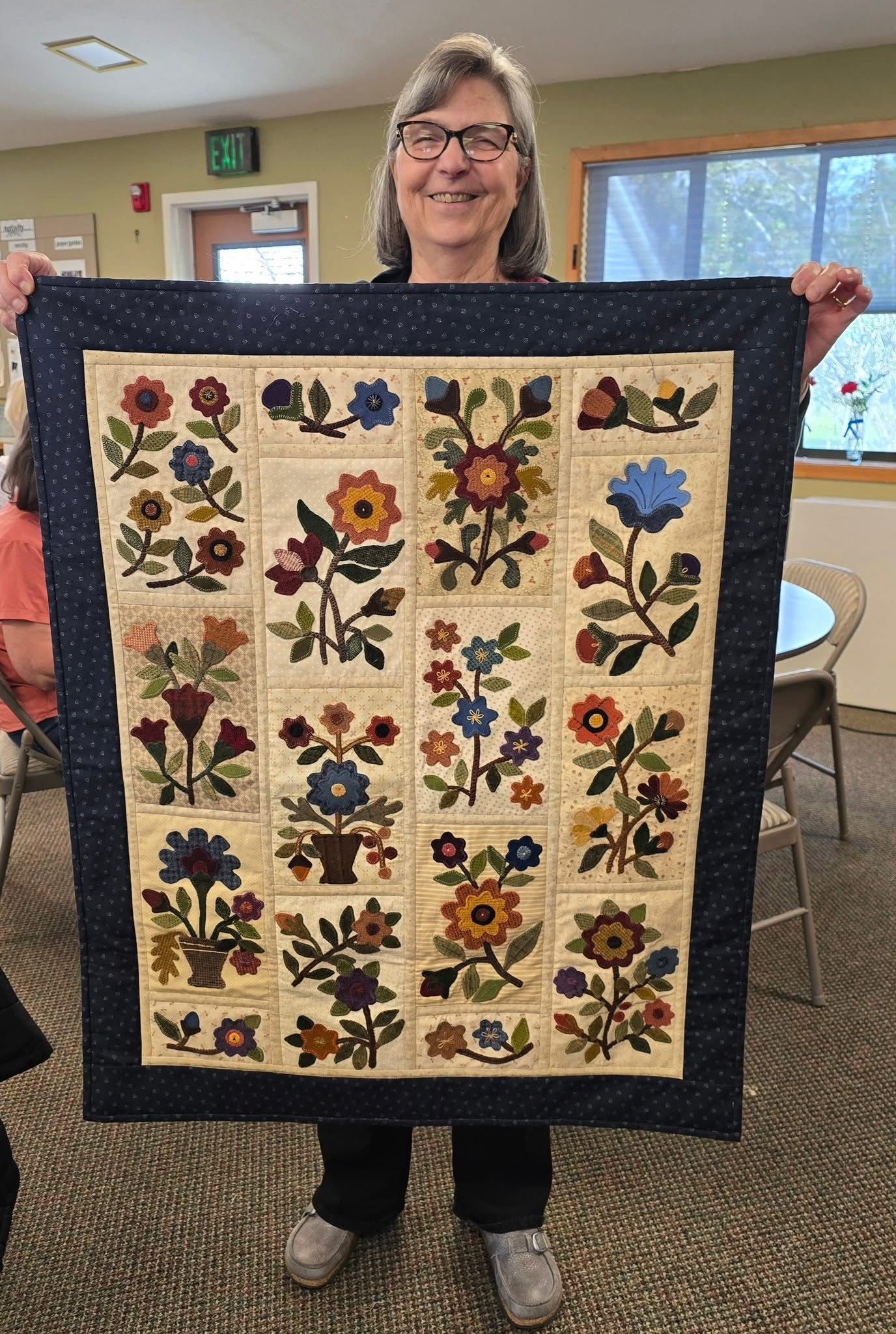 A smiling person holds up a rectangular quilt featuring diverse floral appliqué designs on tan blocks with a dark border.