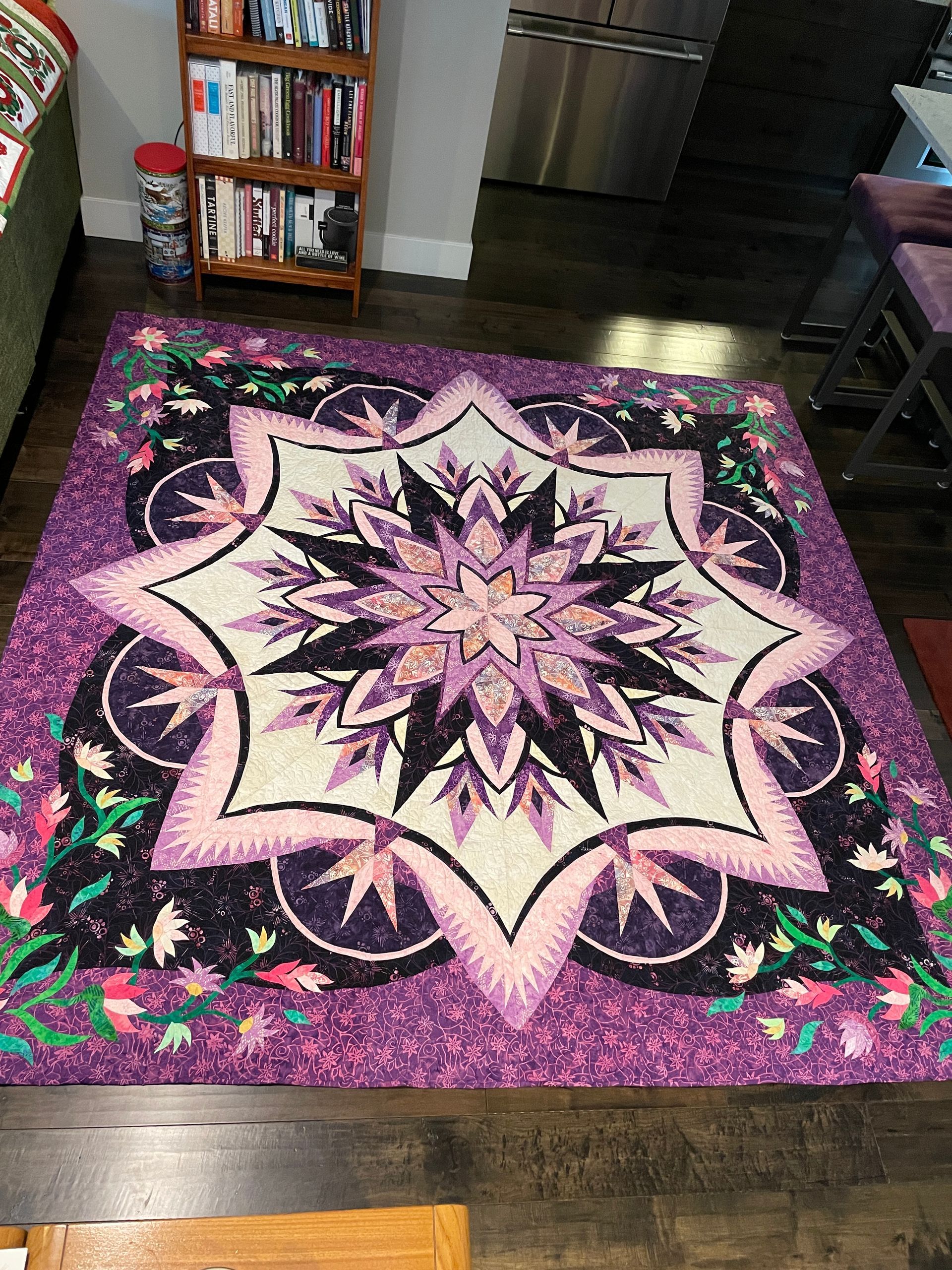 A purple and white star-patterned quilt with floral accents displayed on a dark wood floor near a wooden bookshelf.