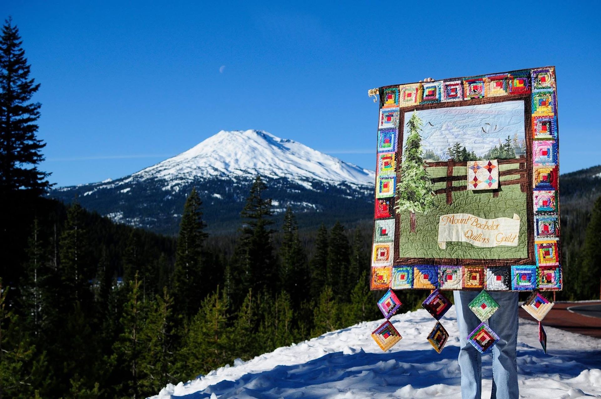 A person holds a colorful, multi-patterned quilt featuring a mountain scene in front of a snow-capped volcanic mountain.