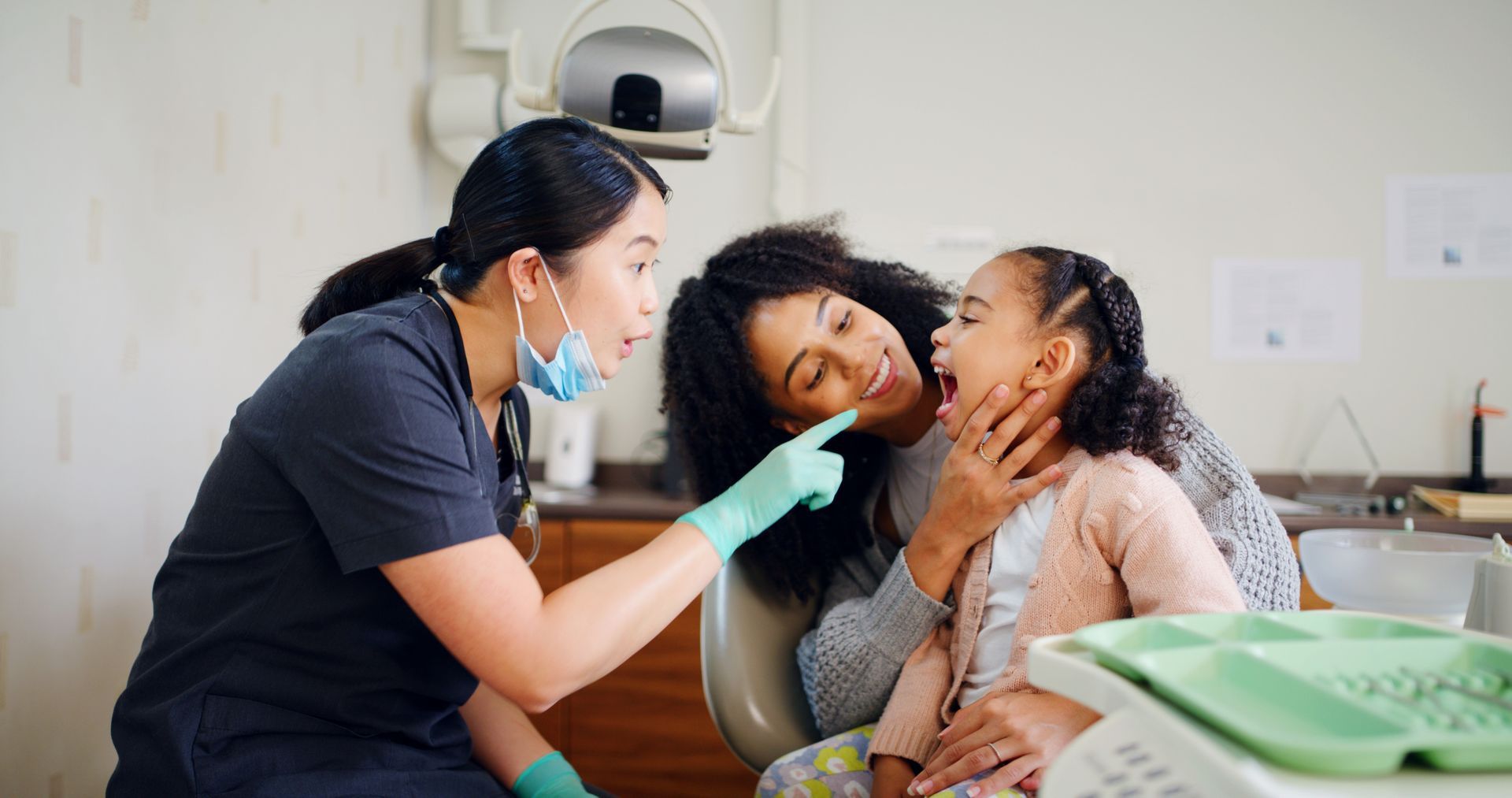 Dentist examining a patient’s mouth while a child sits nearby in a clinic, with green dental tools visible