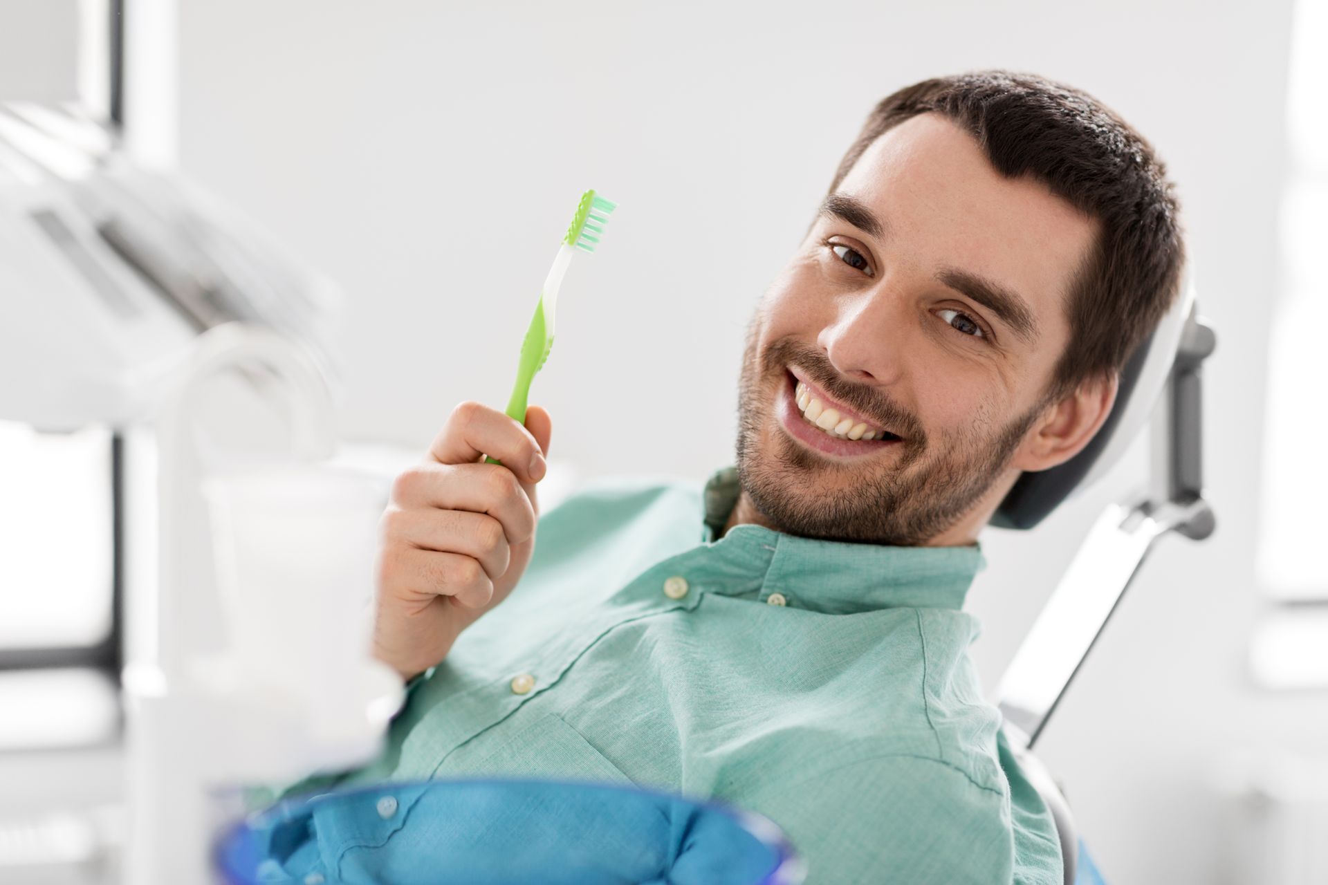 Smiling male dental patient holding a green toothbrush in a dental office chair.