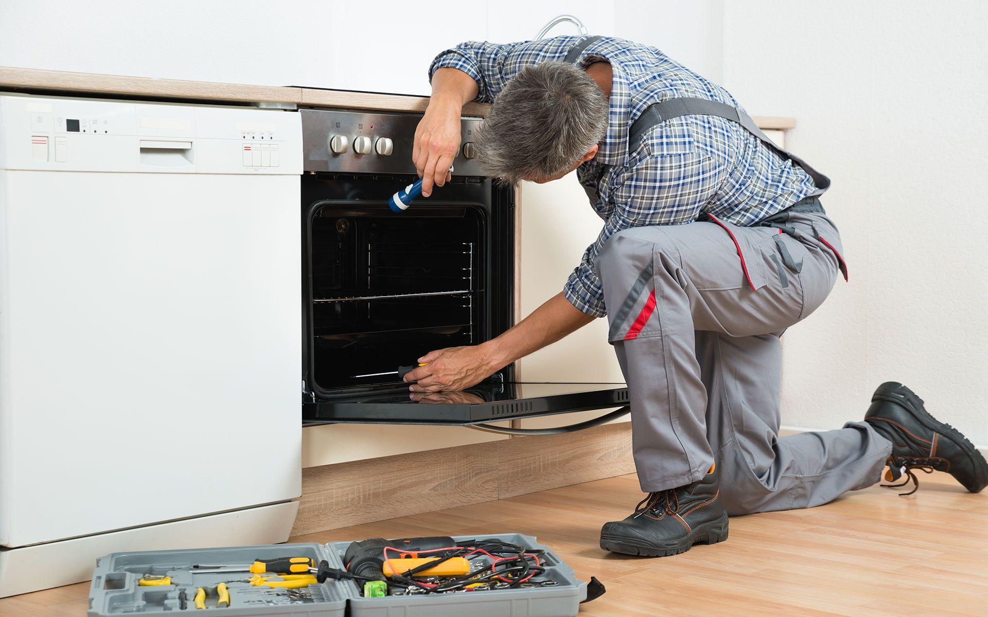 Repairman in grey pants and plaid shirt working on an oven in a kitchen.