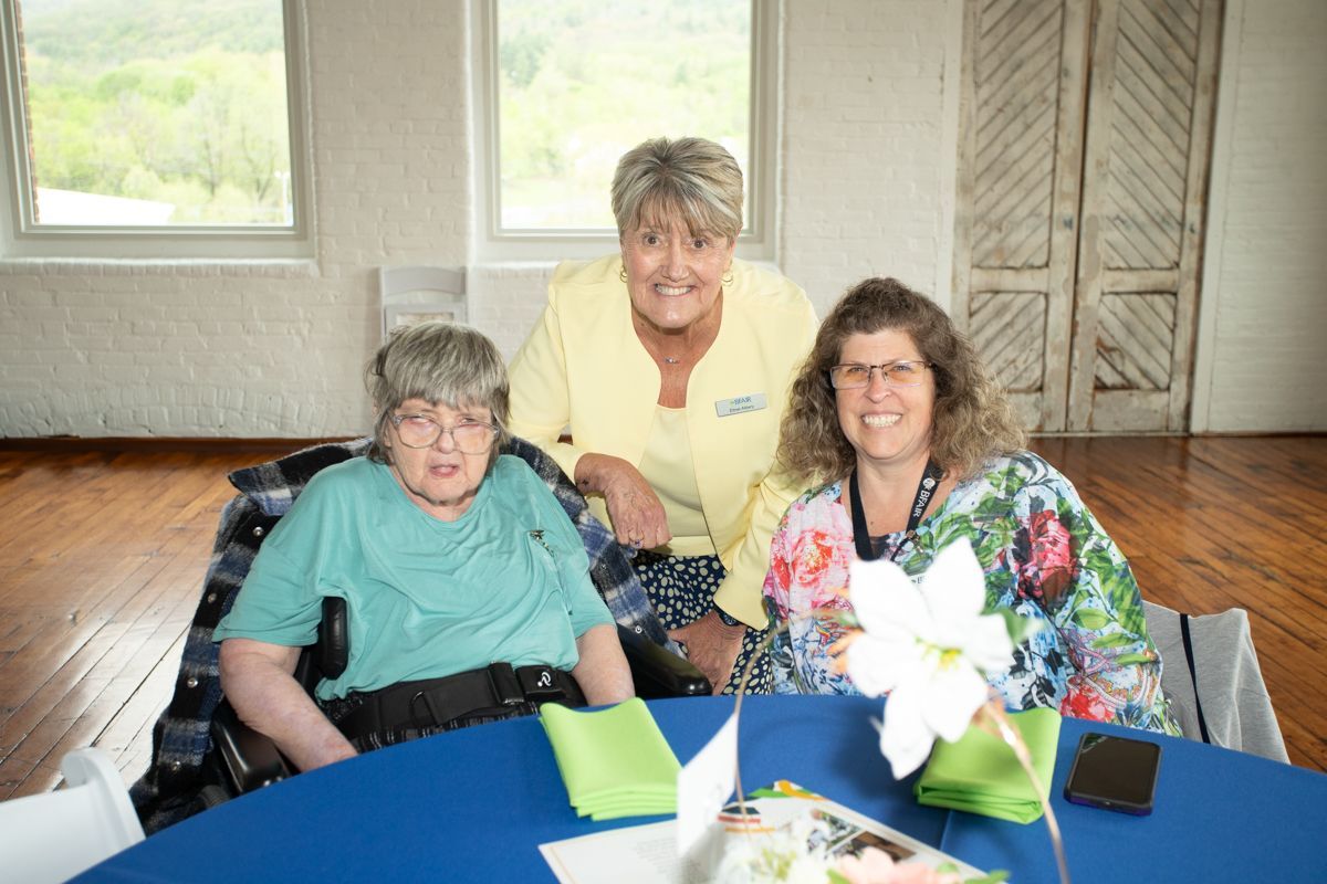 Three women smiling at a table; one in a wheelchair. Brightly colored attire, in a room with windows.