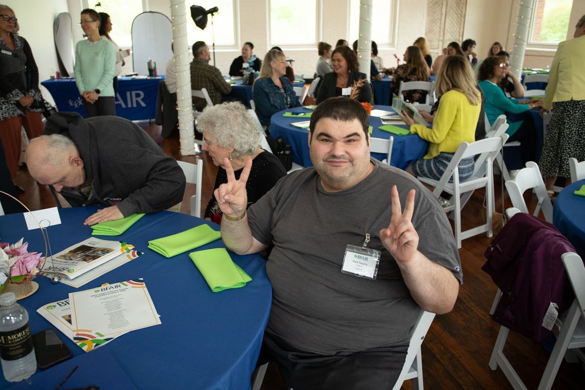 Man at event, smiling, holding up peace sign. People seated at tables, blue tablecloths.