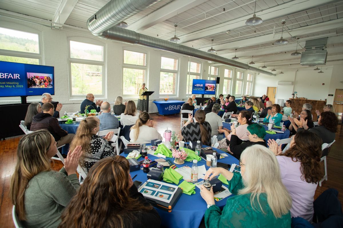 People gathered at tables in a large room applaud as a speaker stands at a podium. Blue tablecloths and two screens.