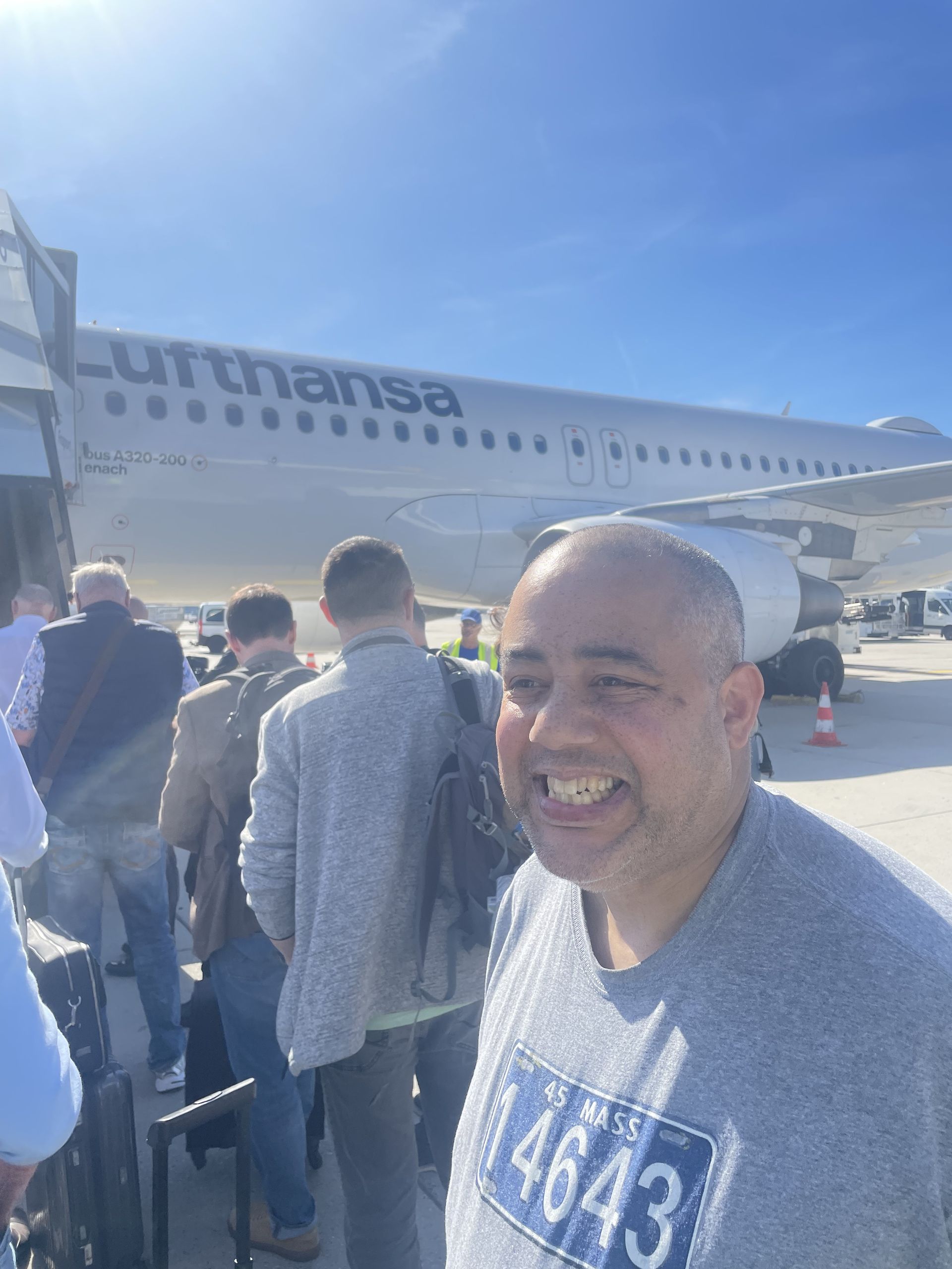Man smiling in front of a Lufthansa plane, boarding with other passengers on a sunny day.