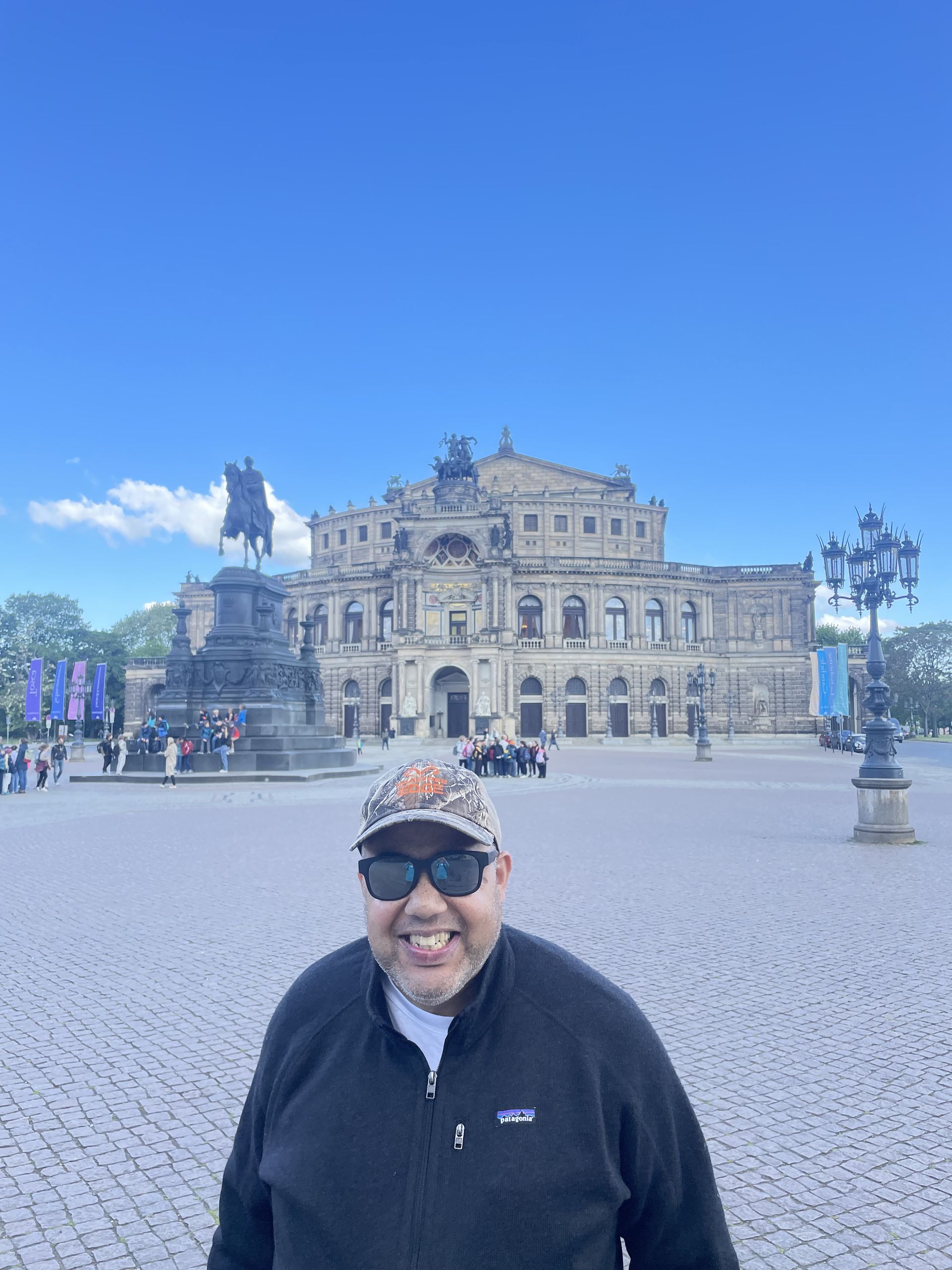 Man smiles in front of the Semper Opera House in Dresden, Germany, blue sky.