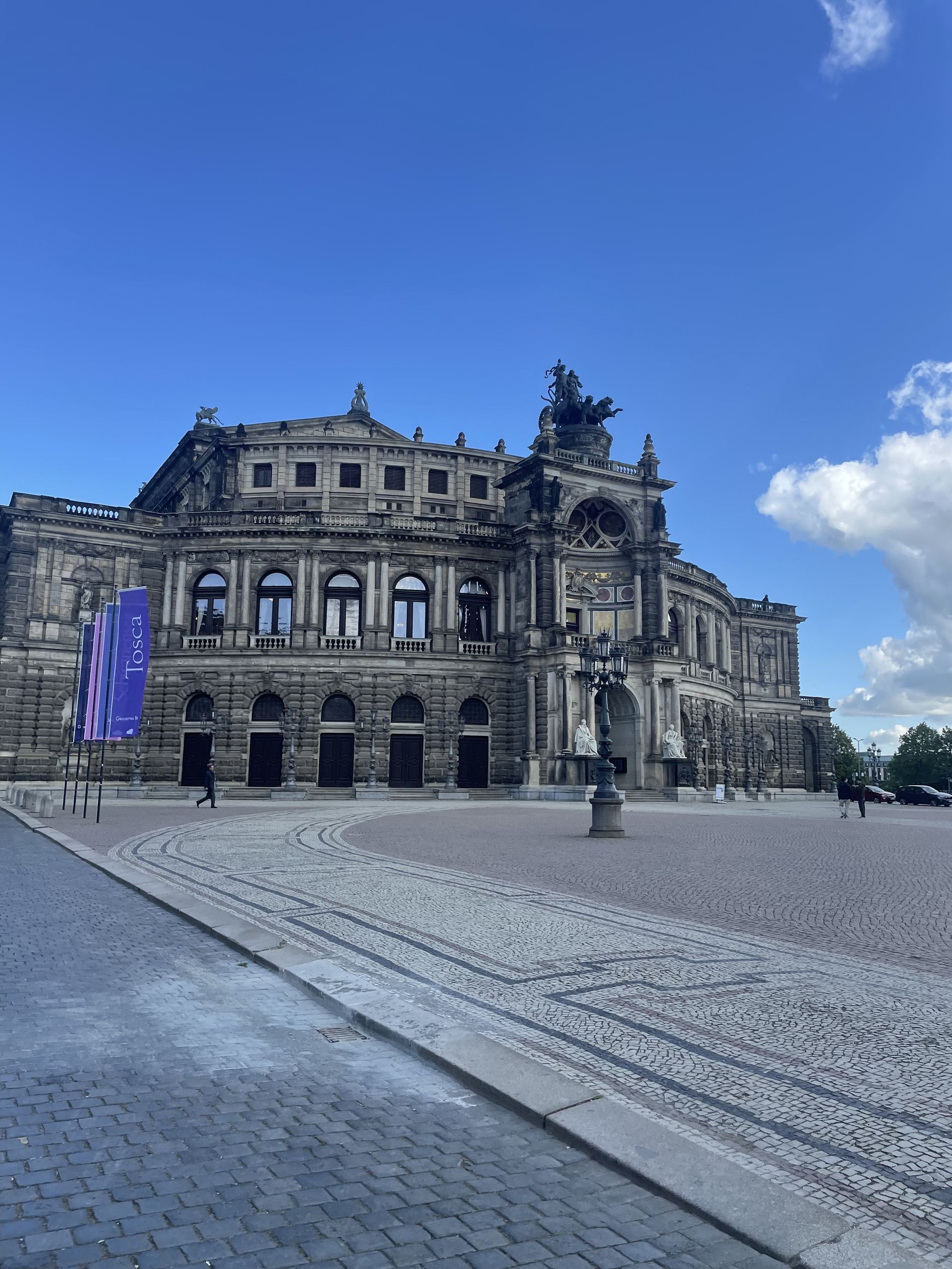 Exterior of the Semper Opera House in Dresden, Germany, on a sunny day. Gray stone building with a paved square in front.