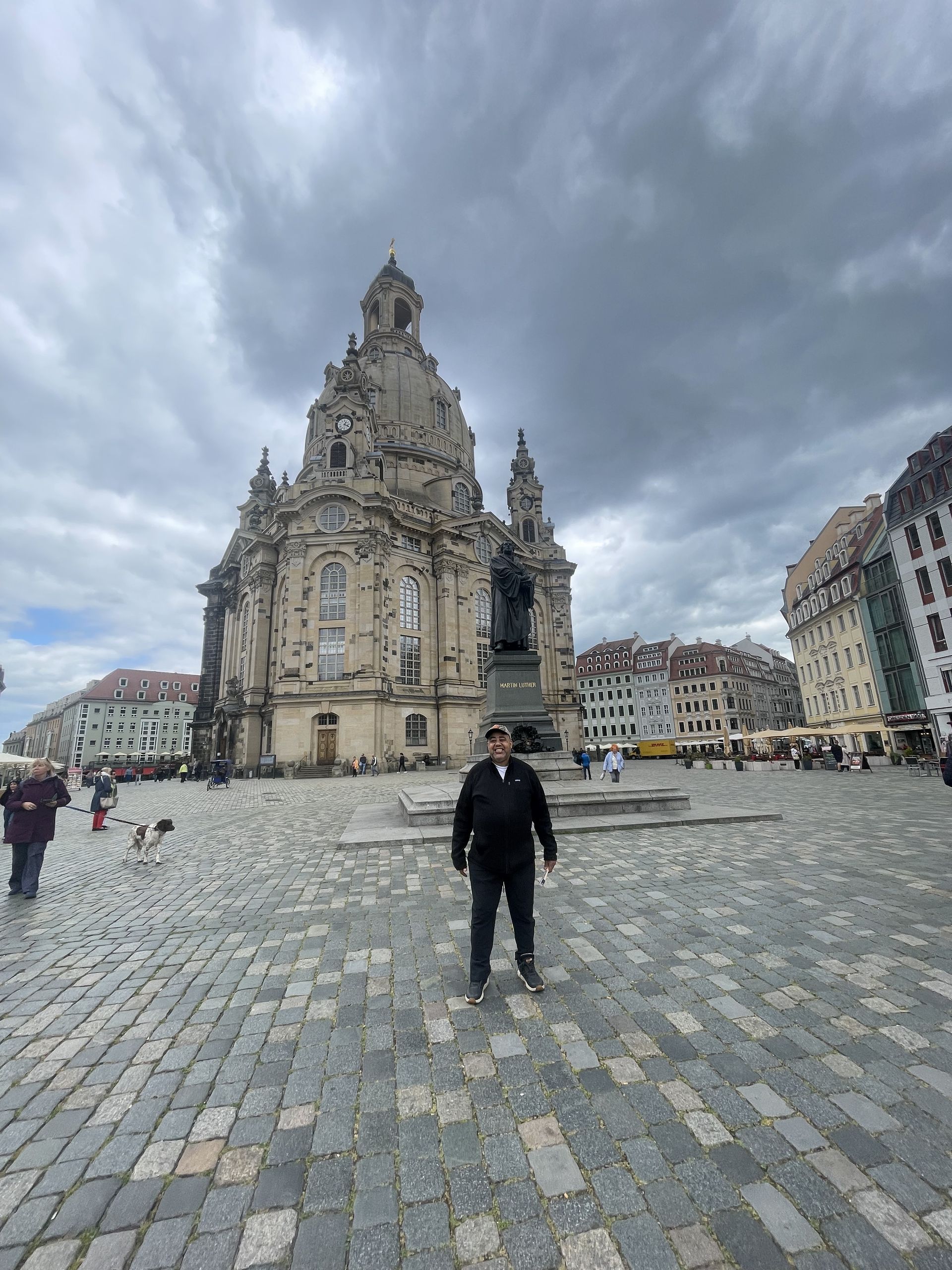 Man stands in a cobblestone square in front of the Frauenkirche church in Dresden, Germany, under a cloudy sky.