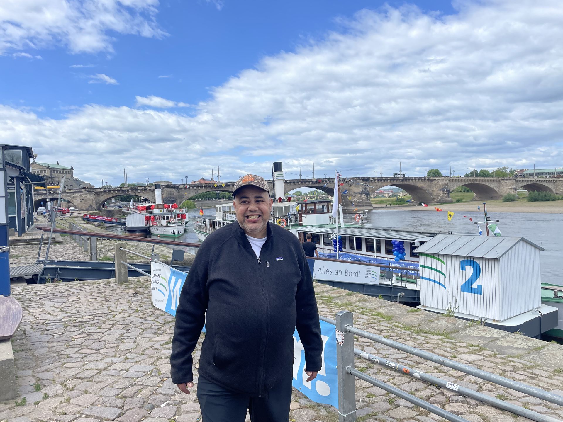 Man smiles at camera on a cobblestone dock with boats, bridge, and cloudy sky.