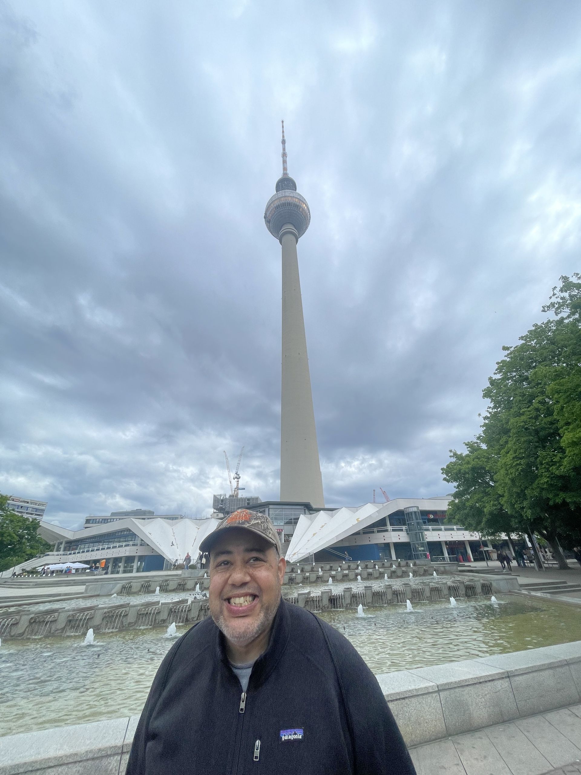 Man smiling in front of the Berlin TV Tower, a tall structure, under a cloudy sky.
