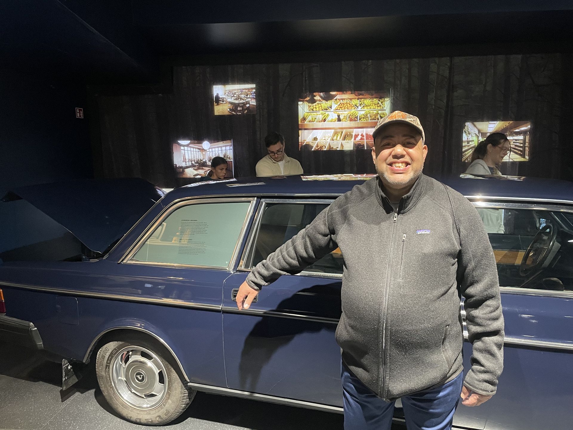 Man in a gray jacket stands next to a blue classic car in a museum.
