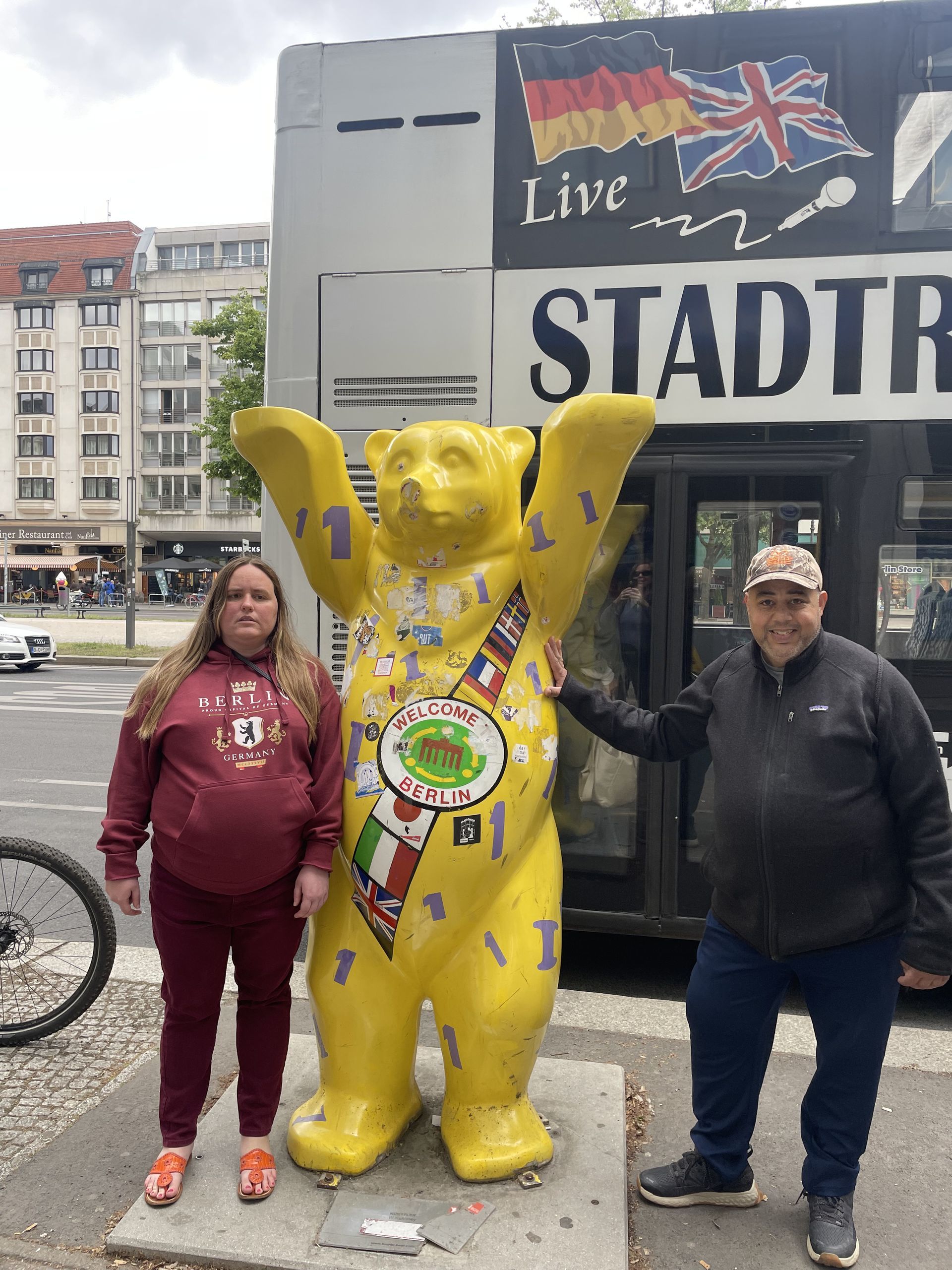 Two people pose with a yellow bear statue in Berlin. A bus with flags is in the background.