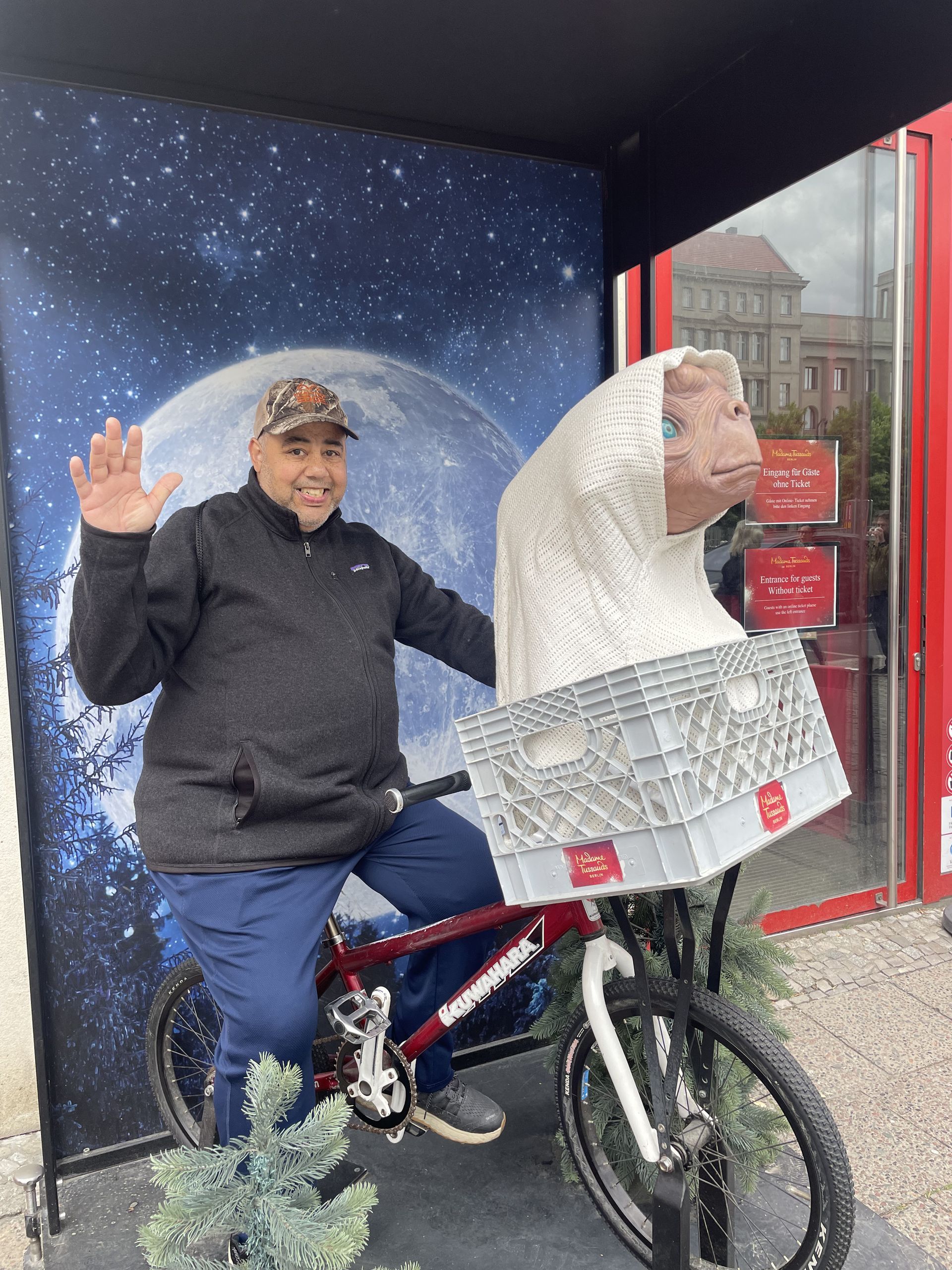 Man on a bike with an E.T. sculpture; moon backdrop, waving, outdoors.