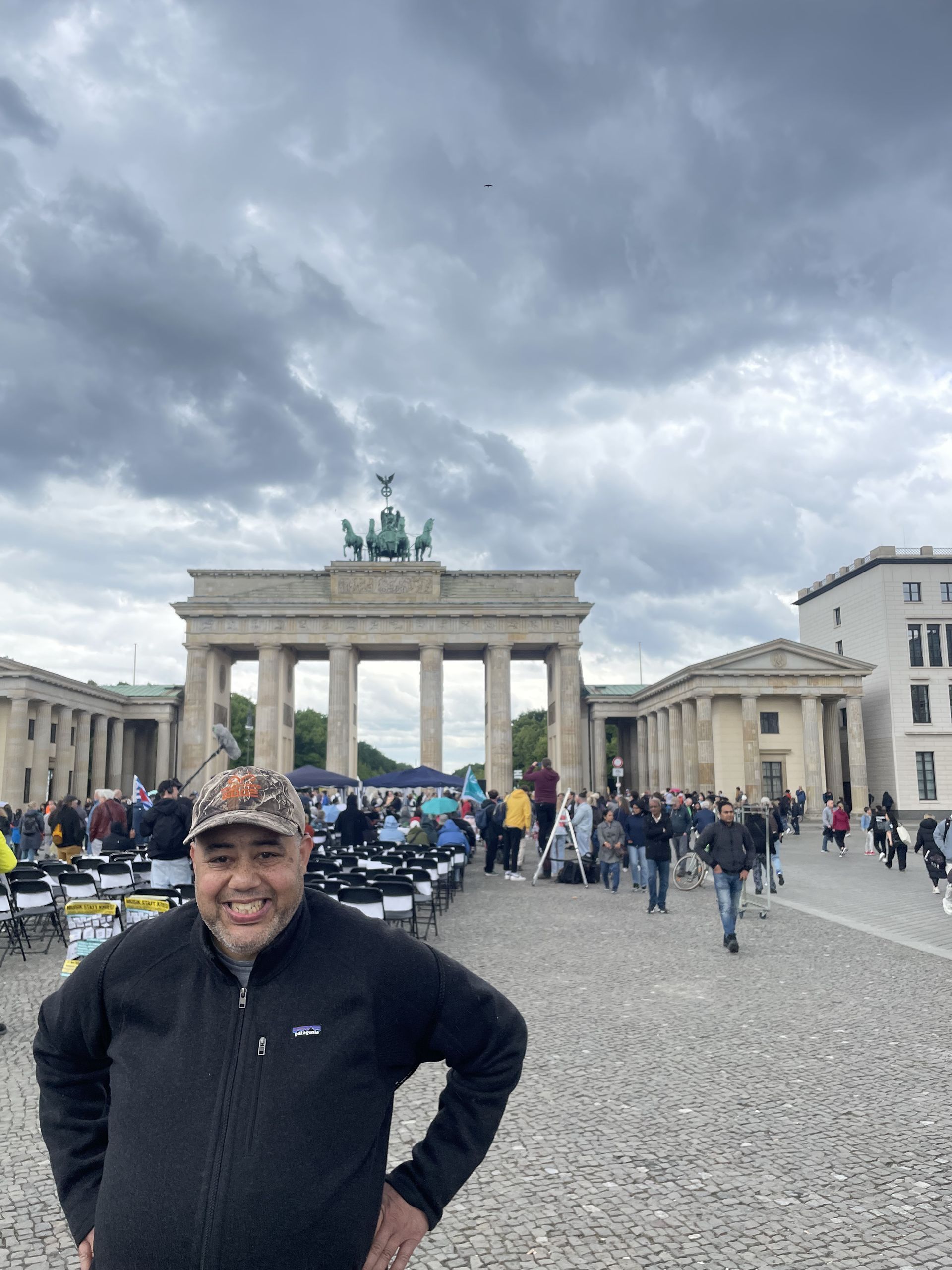 Man smiles in front of Brandenburg Gate, Berlin, cloudy day.