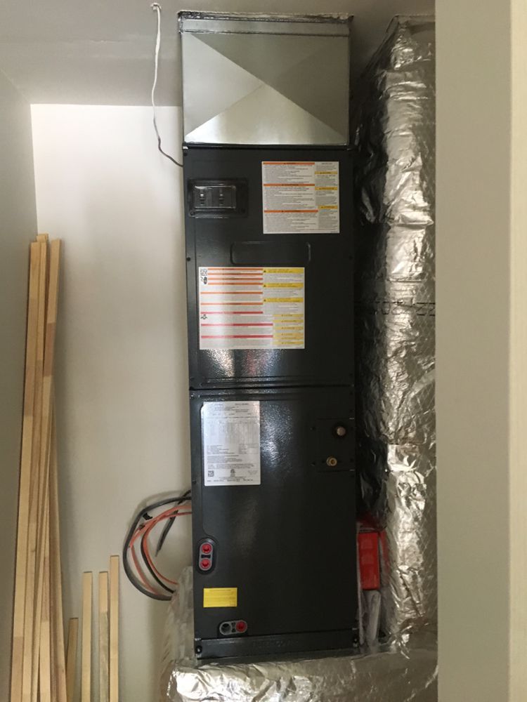 Man in work clothes inspecting an air conditioning unit; indoor setting.