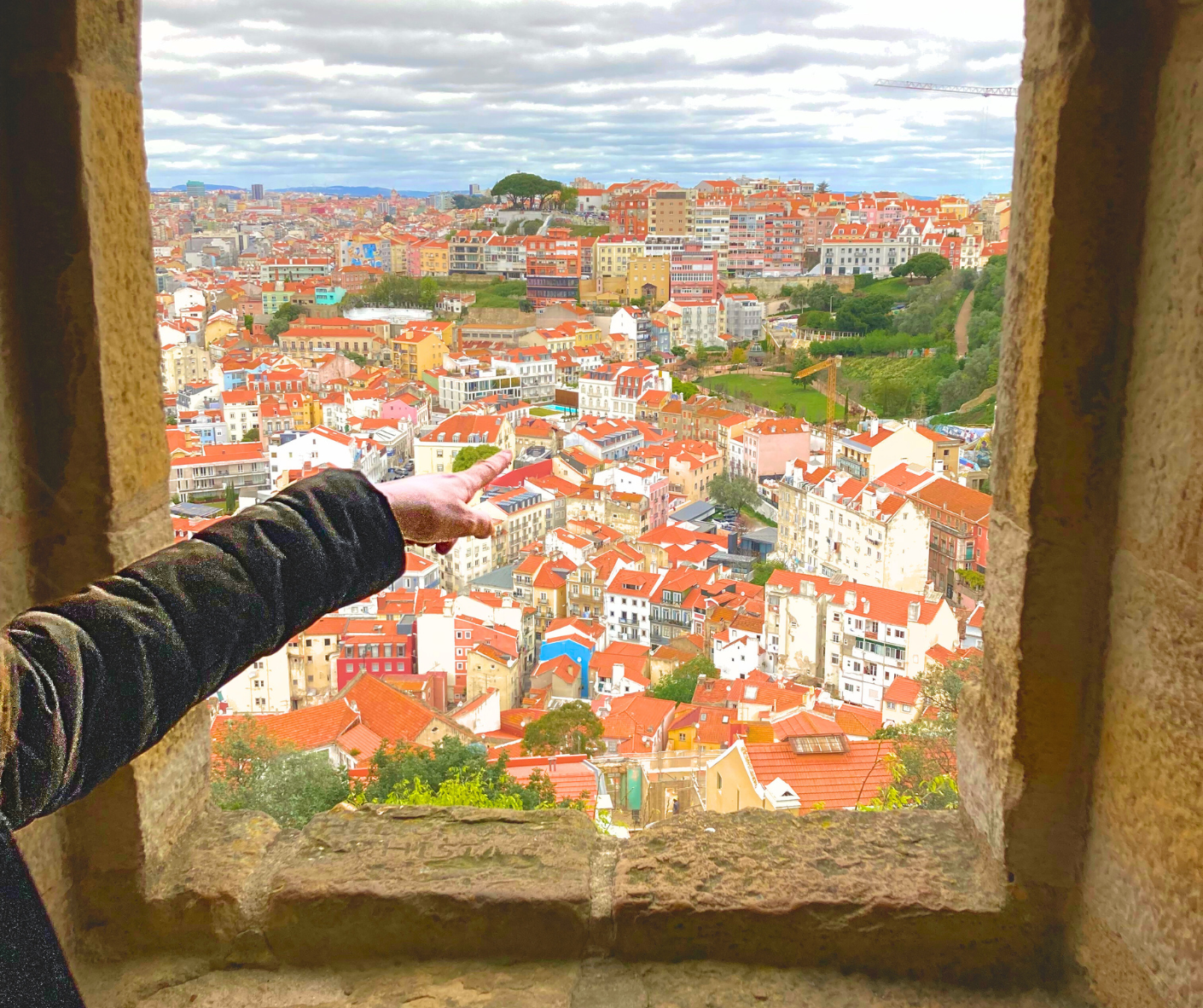 A person points from a stone window toward a dense city landscape filled with buildings featuring red-tiled roofs.