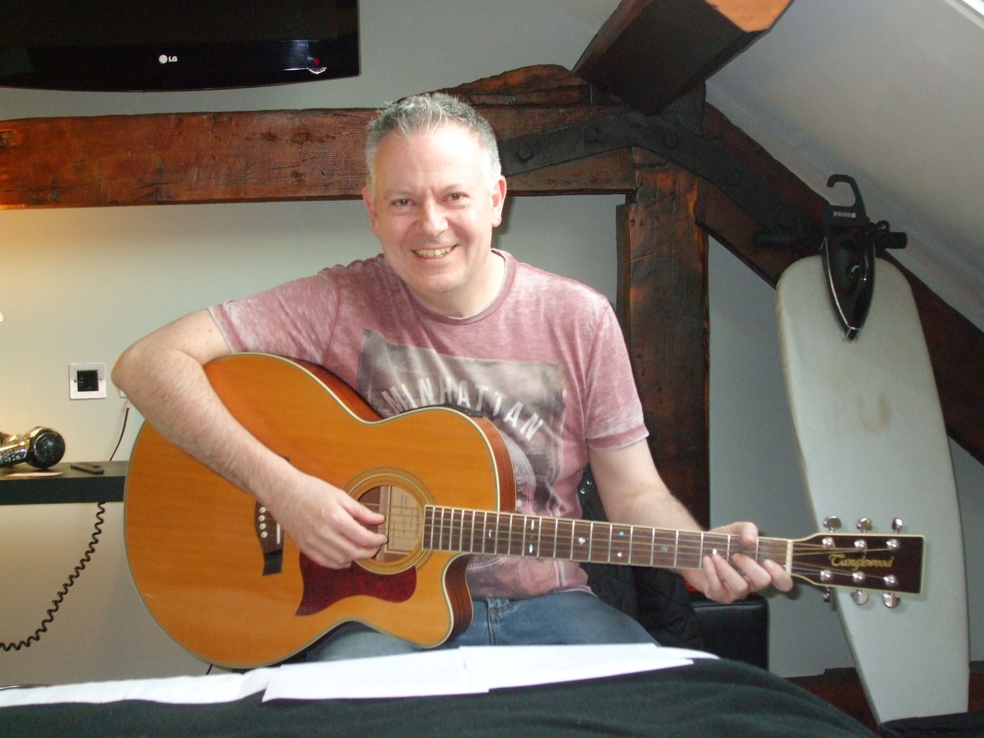 A person with short hair smiles while playing an acoustic guitar in a room with wooden beams and an ironing board nearby.