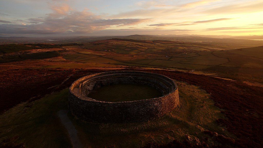 An ancient, circular stone fort sits atop a grassy hill at sunset, overlooking a vast, rolling landscape under a soft sky.
