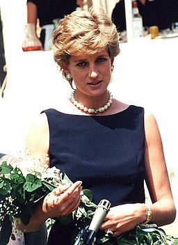 Princess Diana, wearing a dark sleeveless dress and pearl necklace, holding flowers outdoors.