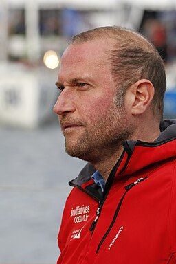 A profile view of a man with a beard, wearing a red jacket, looking toward the left against a blurred harbor background.