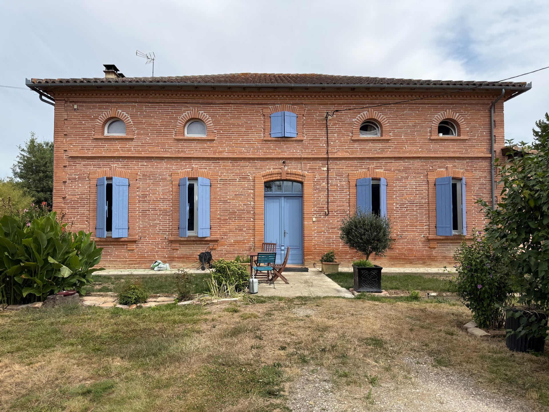 A symmetrical brick farmhouse with blue shutters, a central blue door, and arched attic windows under a tiled roof.