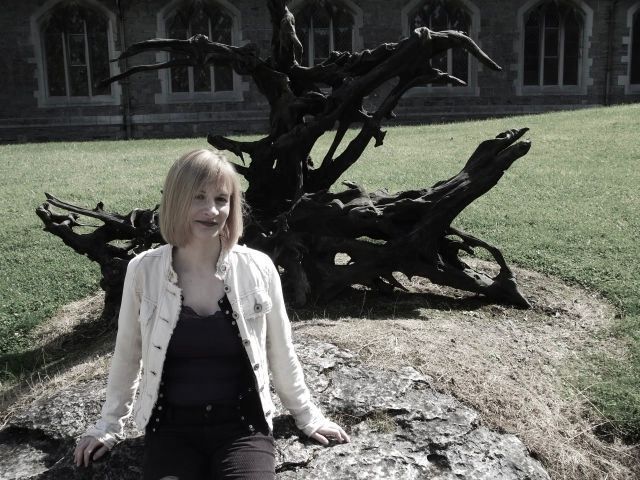 A person in a light jacket sits on a stone surface in front of a large, dark, sculptural tree root on a grassy lawn.