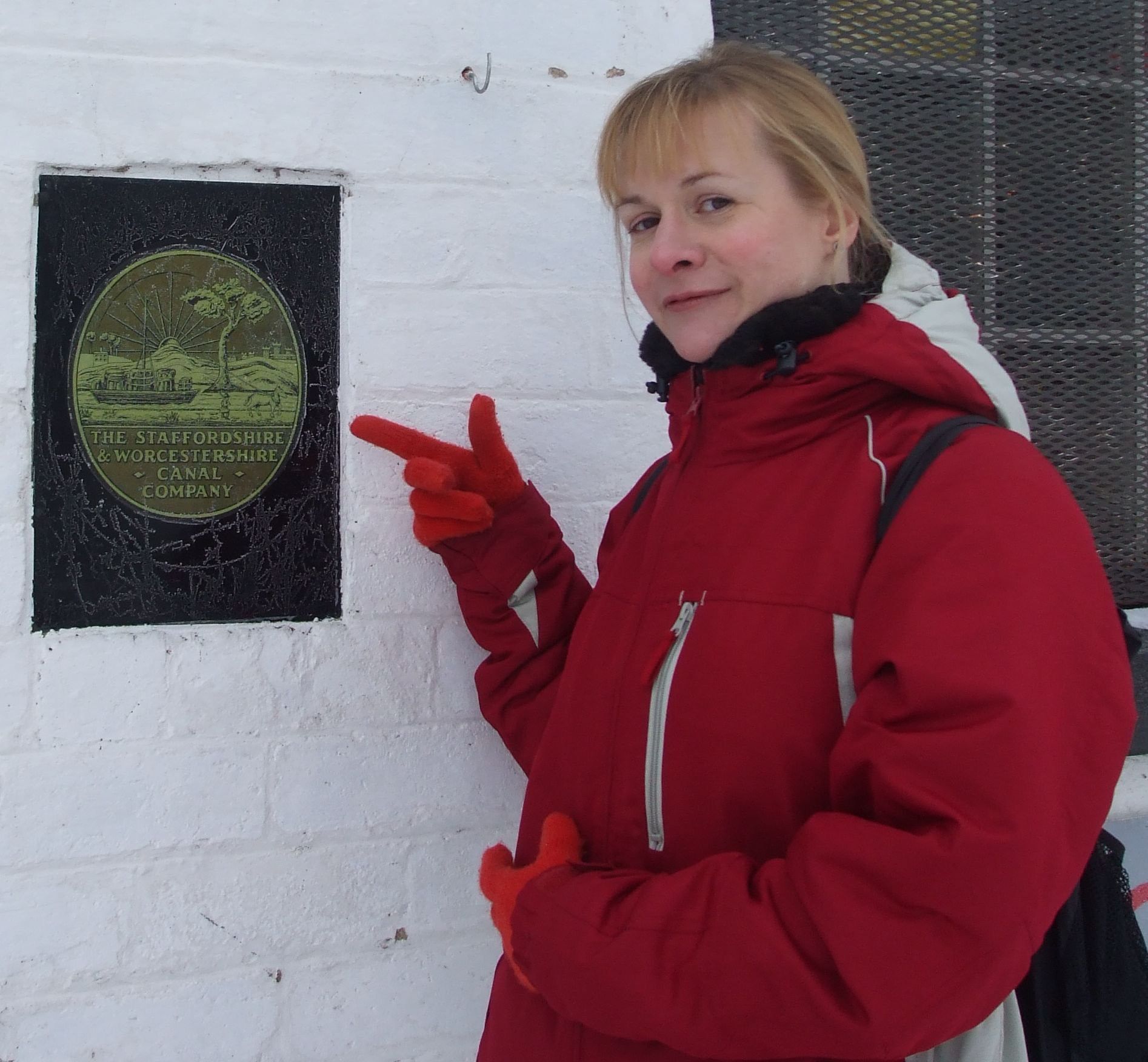 A person in a red coat points to a circular, green commemorative plaque mounted on a white brick wall.