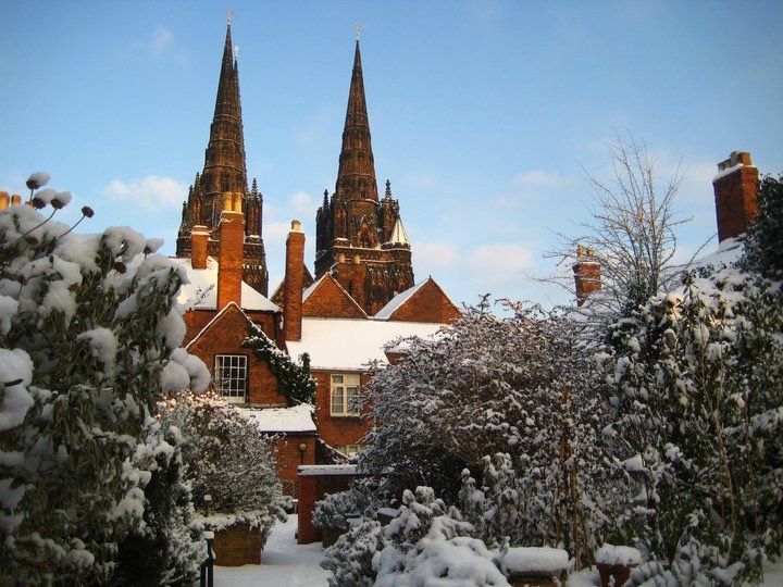 Lichfield Cathedral with twin spires rising above snow-covered gardens and brick buildings on a bright, winter day.