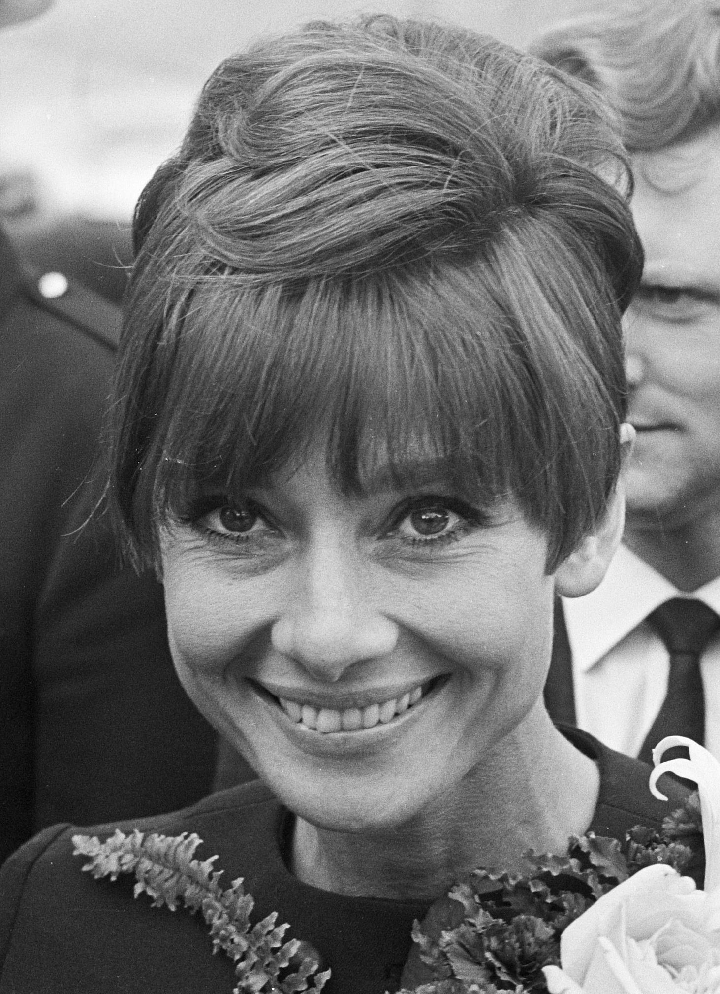 A black-and-white portrait of Audrey Hepburn smiling, featuring her signature beehive hairstyle and bangs.